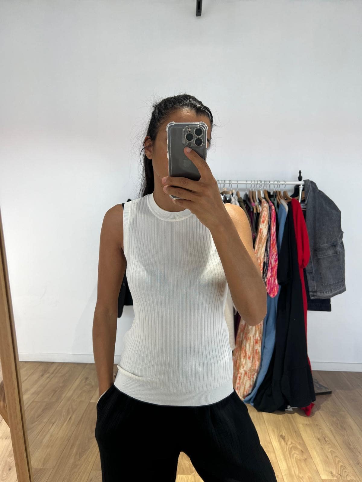 Femme portant un haut sans manche blanc, prenant un selfie devant un miroir avec des vêtements en arrière-plan.