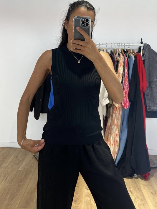 Femme portant un haut sans manche noir, prenant un selfie devant un miroir avec des vêtements en arrière-plan.
