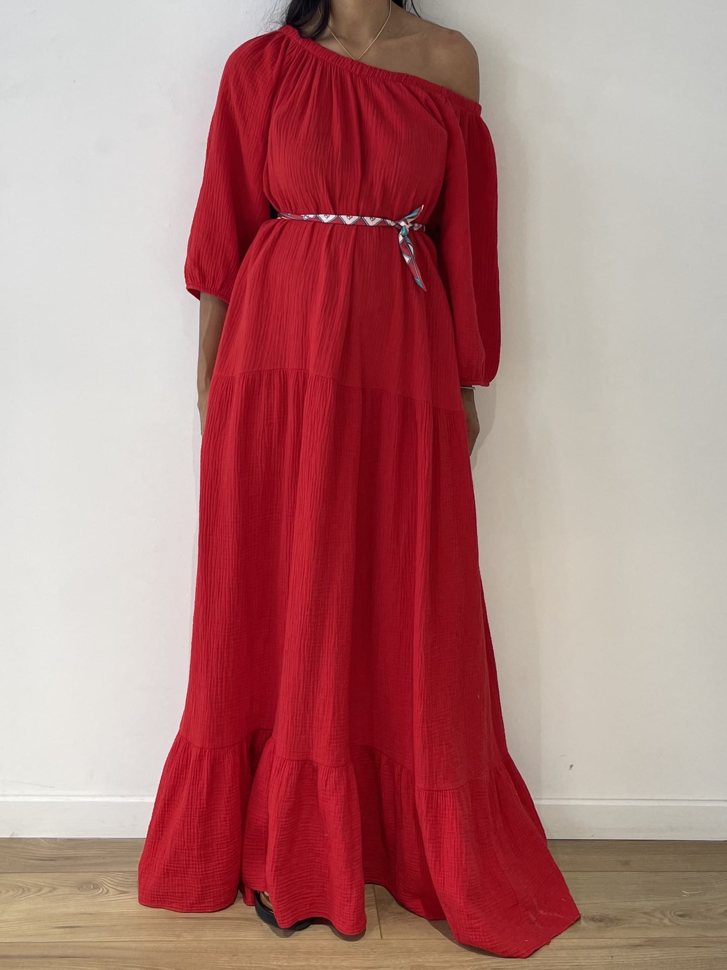 emme portant une robe rouge en gaze de coton avec une épaule dénudée et une ceinture décorative.